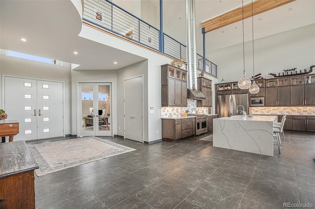 kitchen featuring appliances with stainless steel finishes, a towering ceiling, backsplash, dark brown cabinets, and a center island with sink