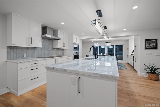 kitchen featuring sink, white cabinets, wall chimney exhaust hood, a center island with sink, and decorative light fixtures