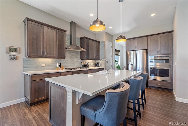 kitchen with wall chimney range hood, a kitchen island with sink, pendant lighting, and appliances with stainless steel finishes