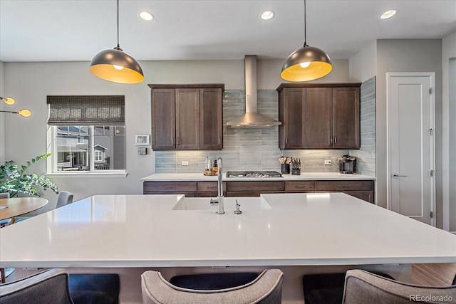 kitchen with wall chimney exhaust hood, pendant lighting, a kitchen island with sink, stainless steel gas cooktop, and backsplash