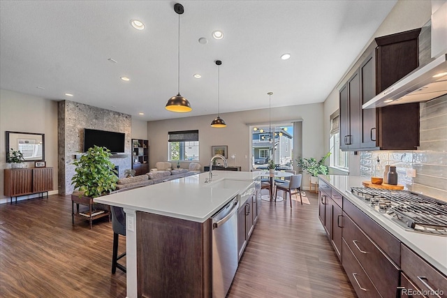 kitchen with wall chimney exhaust hood, decorative light fixtures, stainless steel appliances, tasteful backsplash, and a kitchen island with sink