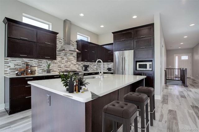 kitchen with wall chimney range hood, a center island with sink, appliances with stainless steel finishes, light wood-type flooring, and sink