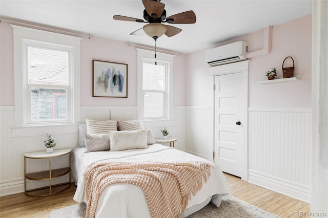 bedroom with a wall mounted air conditioner, light wood finished floors, a ceiling fan, and a wainscoted wall
