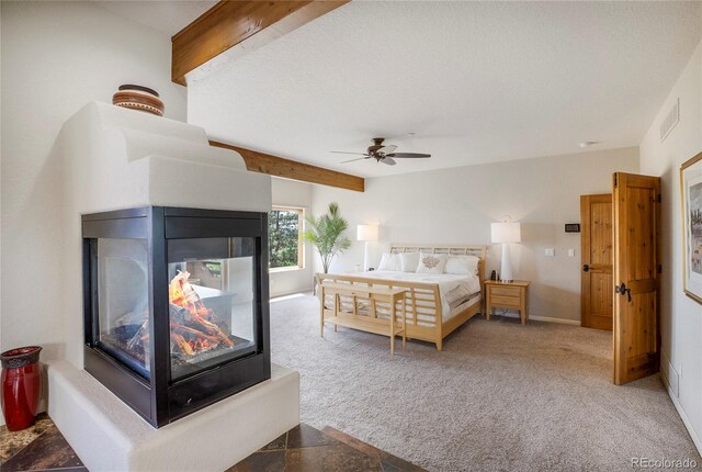 bedroom with ceiling fan, a multi sided fireplace, carpet flooring, a textured ceiling, and beamed ceiling