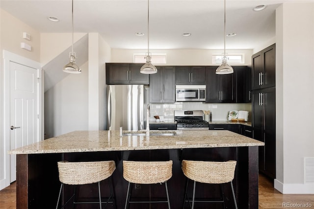 kitchen with stainless steel appliances, light stone countertops, a center island with sink, and wood finished floors