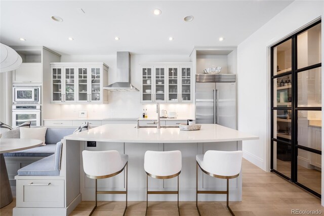 kitchen featuring wall chimney range hood, built in appliances, a sink, light wood-style floors, and a breakfast bar