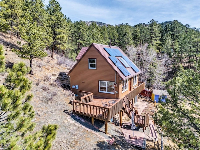 back of house featuring metal roof, a view of trees, roof mounted solar panels, and a wooden deck