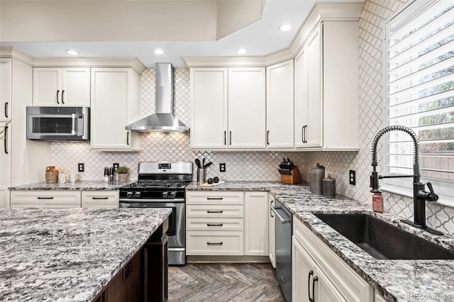 kitchen featuring wall chimney exhaust hood, appliances with stainless steel finishes, a sink, backsplash, and light stone counters