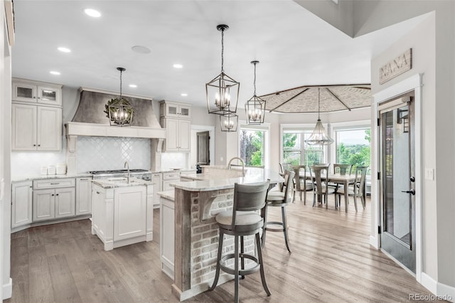 kitchen featuring premium range hood, decorative light fixtures, an island with sink, and light hardwood / wood-style flooring