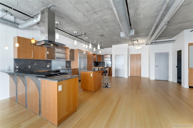 kitchen featuring brown cabinetry, island range hood, light wood-type flooring, stainless steel built in refrigerator, and a kitchen bar