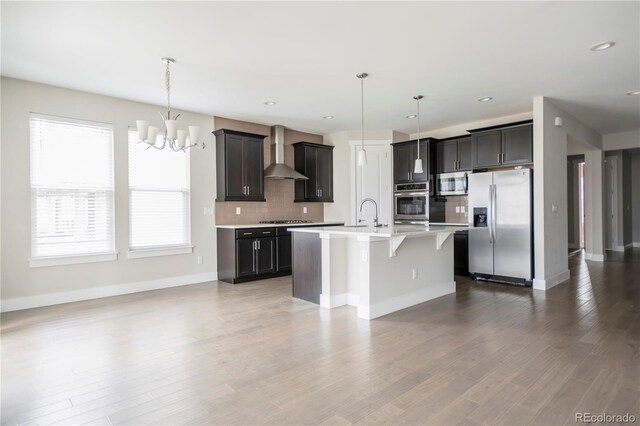 kitchen featuring decorative backsplash, light countertops, wall chimney exhaust hood, a notable chandelier, and stainless steel appliances