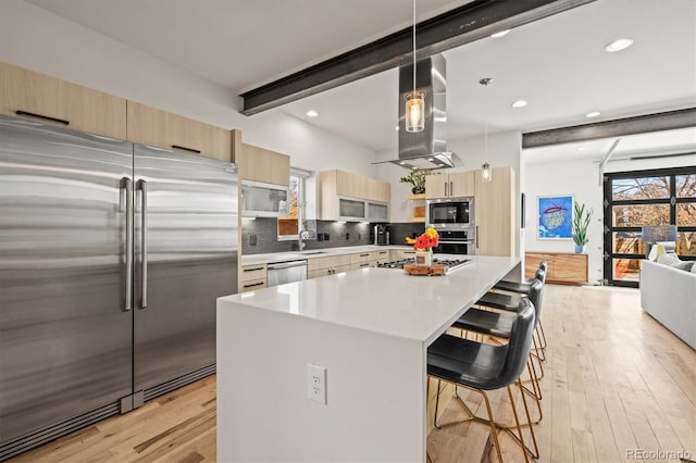 kitchen with beam ceiling, light brown cabinetry, ventilation hood, appliances with stainless steel finishes, and a breakfast bar area