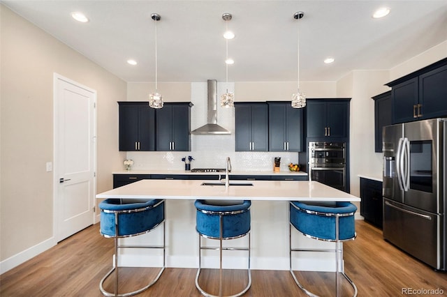 kitchen featuring light wood-style floors, appliances with stainless steel finishes, a kitchen island with sink, wall chimney range hood, and a kitchen bar