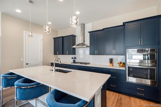 kitchen with stainless steel appliances, a sink, wall chimney exhaust hood, light wood finished floors, and a kitchen bar