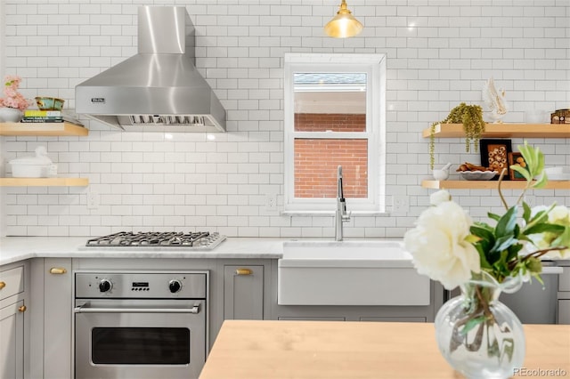 kitchen featuring wall chimney exhaust hood, stainless steel appliances, a sink, open shelves, and gray cabinets