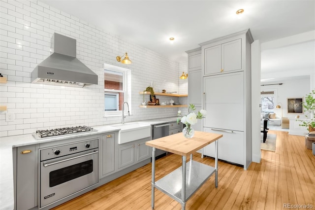 kitchen with a sink, wall chimney exhaust hood, appliances with stainless steel finishes, gray cabinetry, and light wood-type flooring