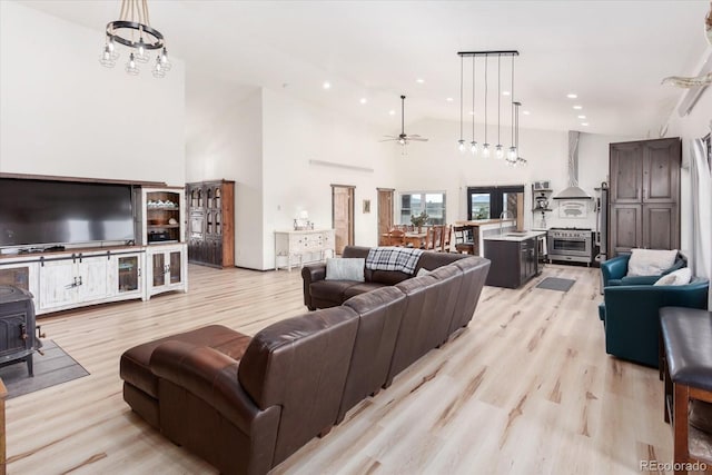 living room with sink, a towering ceiling, light hardwood / wood-style floors, ceiling fan with notable chandelier, and a wood stove