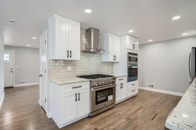 kitchen with light stone counters, backsplash, light wood-style floors, appliances with stainless steel finishes, and wall chimney exhaust hood