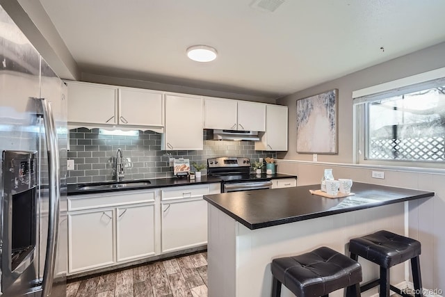 kitchen with dark countertops, appliances with stainless steel finishes, a breakfast bar, under cabinet range hood, and a sink