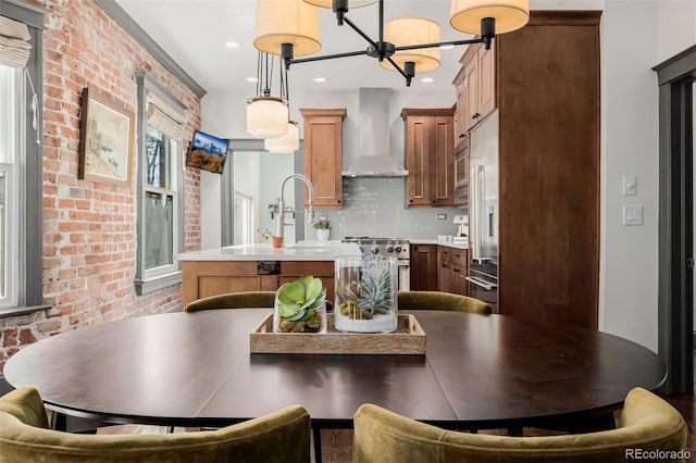 kitchen with a center island with sink, brick wall, a sink, light countertops, and wall chimney range hood