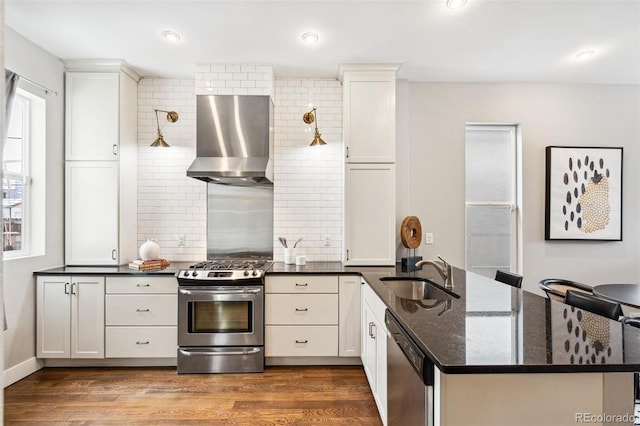 kitchen featuring appliances with stainless steel finishes, wall chimney exhaust hood, white cabinetry, dark stone counters, and sink