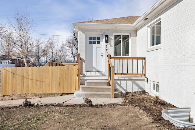 view of exterior entry with roof with shingles, fence, and brick siding