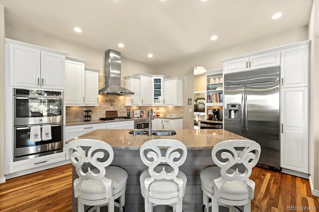 kitchen with wall chimney exhaust hood, dark wood-type flooring, stainless steel appliances, light stone counters, and a center island with sink