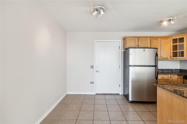 kitchen with dark stone countertops, freestanding refrigerator, light tile patterned floors, glass insert cabinets, and baseboards