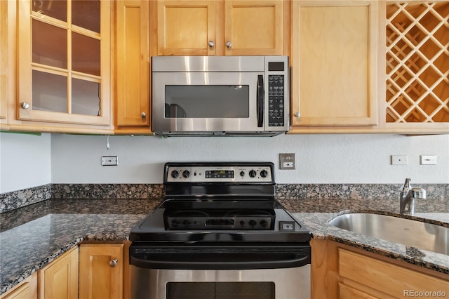 kitchen with light brown cabinetry, a sink, stainless steel appliances, dark stone counters, and glass insert cabinets