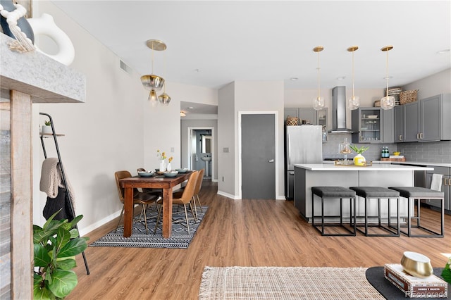 kitchen with stainless steel refrigerator, decorative light fixtures, gray cabinetry, a kitchen bar, and wall chimney exhaust hood