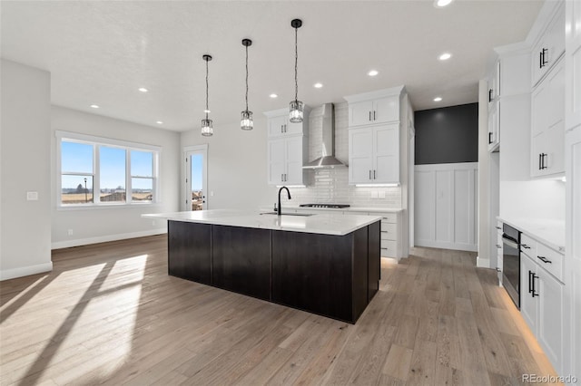 kitchen featuring a kitchen island with sink, wall chimney range hood, white cabinetry, and pendant lighting