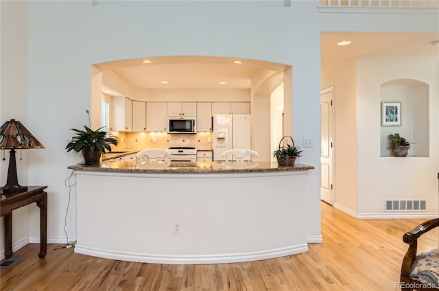 kitchen featuring white cabinetry, light hardwood / wood-style flooring, kitchen peninsula, dark stone counters, and white appliances