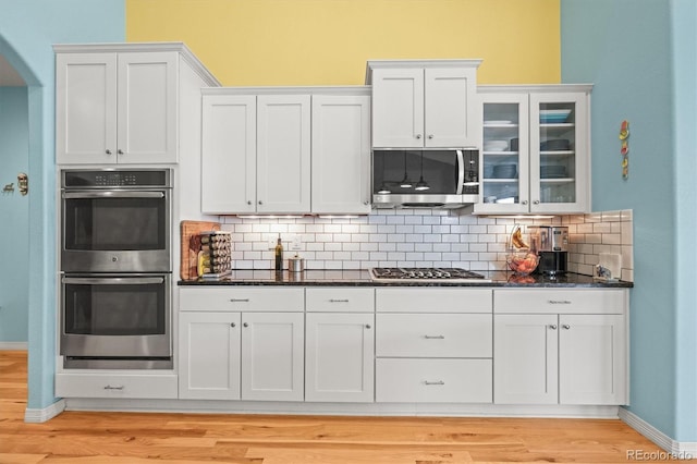 kitchen with tasteful backsplash, glass insert cabinets, light wood-type flooring, dark stone countertops, and appliances with stainless steel finishes