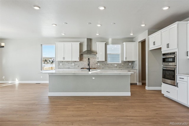 kitchen with white cabinetry, light wood-type flooring, an island with sink, stainless steel double oven, and wall chimney range hood