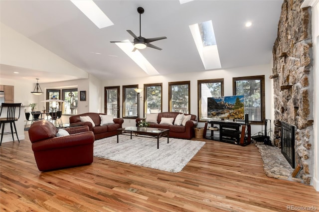 living room featuring high vaulted ceiling, a stone fireplace, a skylight, a ceiling fan, and light wood finished floors