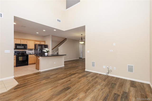 kitchen with light brown cabinetry, hanging light fixtures, light wood-type flooring, a kitchen island with sink, and black appliances