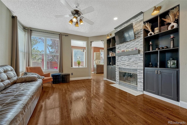 living room featuring built in features, a textured ceiling, wood finished floors, a stone fireplace, and ceiling fan