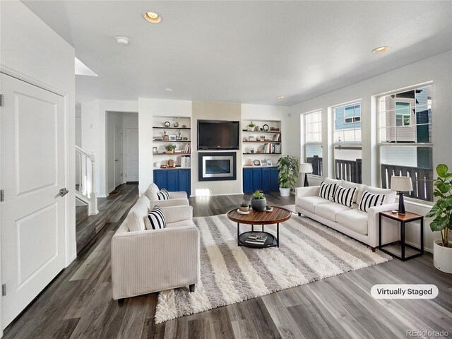 living room with a glass covered fireplace, built in shelves, stairs, and dark wood-type flooring