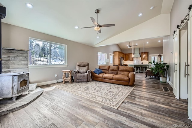 living room with wood finished floors, a barn door, ceiling fan, a wood stove, and vaulted ceiling