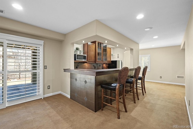 kitchen with brown cabinets, light colored carpet, stainless steel microwave, and a breakfast bar