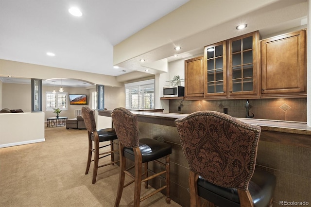 kitchen featuring arched walkways, stainless steel microwave, decorative backsplash, brown cabinetry, and a kitchen breakfast bar