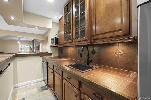 kitchen with brown cabinetry, glass insert cabinets, appliances with stainless steel finishes, a sink, and backsplash