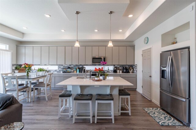 kitchen with stainless steel appliances, hanging light fixtures, a center island with sink, a raised ceiling, and hardwood / wood-style floors