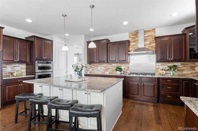 kitchen featuring stainless steel appliances, dark wood-type flooring, a kitchen island, wall chimney range hood, and a kitchen bar