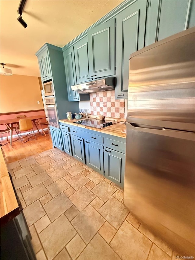 kitchen featuring track lighting, under cabinet range hood, stainless steel appliances, butcher block countertops, and backsplash