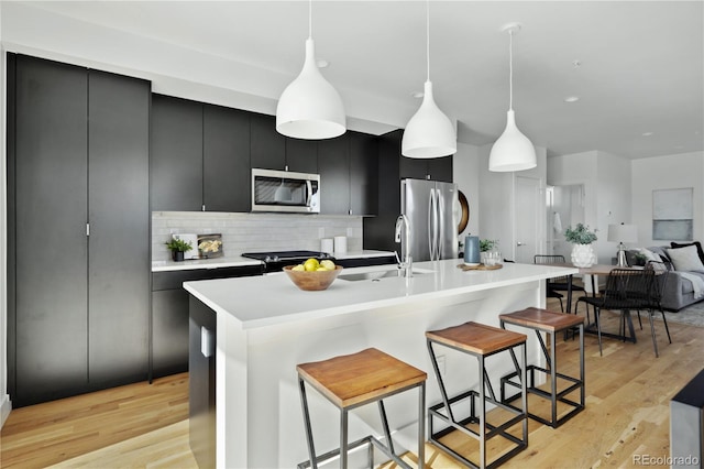 kitchen with appliances with stainless steel finishes, an island with sink, sink, hanging light fixtures, and light hardwood / wood-style flooring