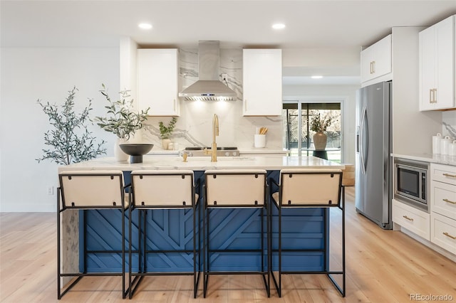 kitchen featuring a kitchen bar, appliances with stainless steel finishes, light wood-type flooring, wall chimney exhaust hood, and white cabinetry