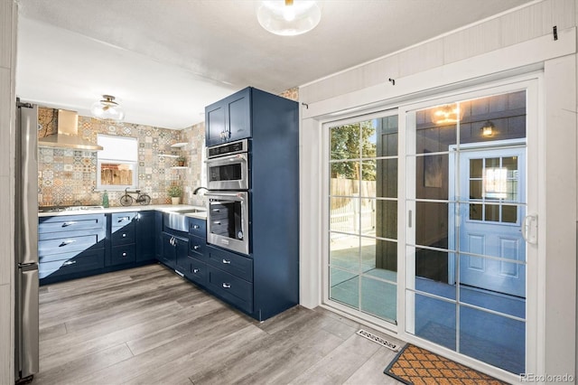 kitchen with wall chimney exhaust hood, blue cabinets, tasteful backsplash, light wood-type flooring, and appliances with stainless steel finishes