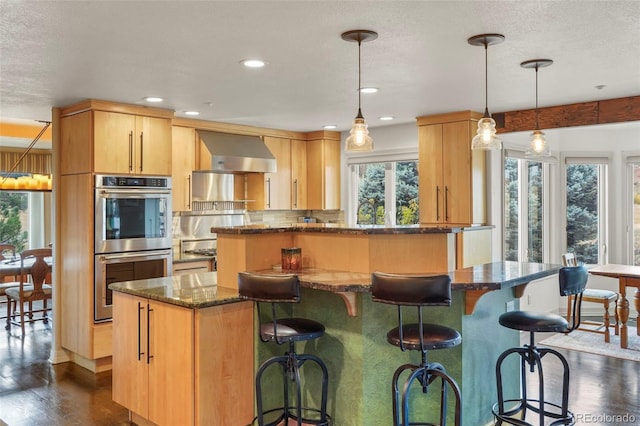 kitchen featuring double oven, light brown cabinets, wall chimney range hood, dark hardwood / wood-style floors, and pendant lighting