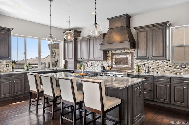 10468 Ladera Dr, Lone Tree, CO 80124 - kitchen featuring custom exhaust hood, a center island with sink, backsplash, dark brown cabinets, and light stone counters